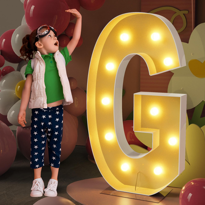 Child standing next to a large illuminated letter 'G' with colorful balloons in the background