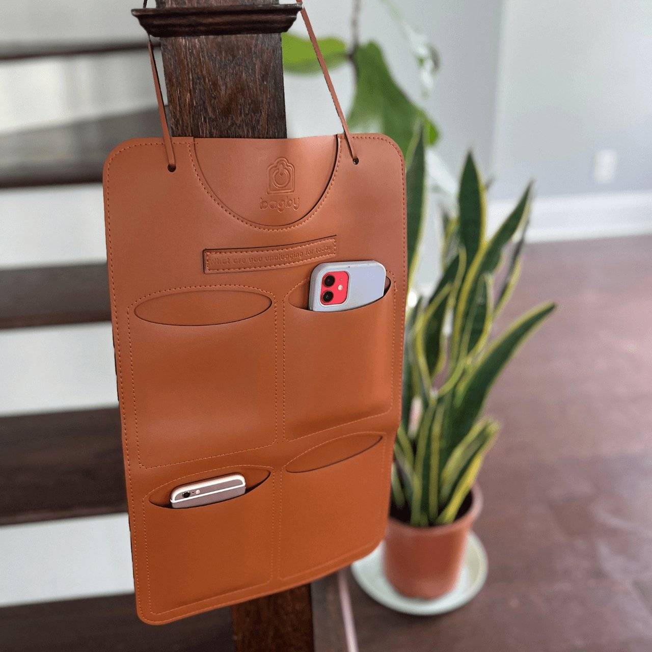 Brown leather phone holder with two phone cases attached, hanging on a wooden post with a plant in the background.