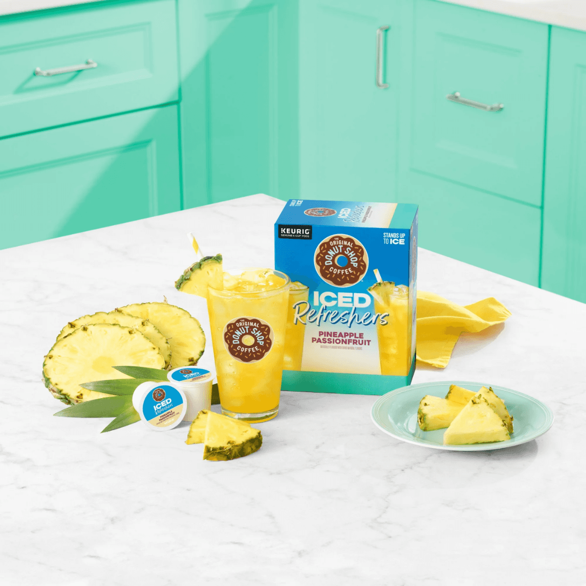 Tropical kitchen scene with Pineapple Passionfruit K-Cup Iced Refreshers box, fresh pineapple slices, and a glass of iced drink on a counter.