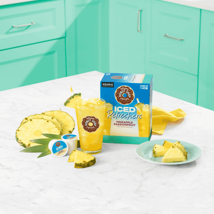 Tropical kitchen scene with Pineapple Passionfruit K-Cup Iced Refreshers box, fresh pineapple slices, and a glass of iced drink on a counter.