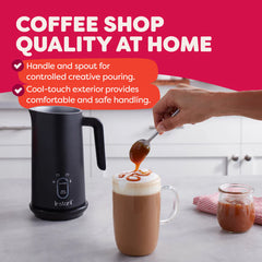 Black milk frother with a hand pouring syrup into a coffee cup on a kitchen counter, against a pink background with text about product features.
