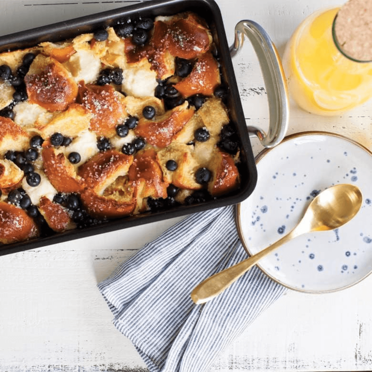 Baked dish with bread and blueberries in a Nordic Ware baking pan on a white surface with a plate and spoon.