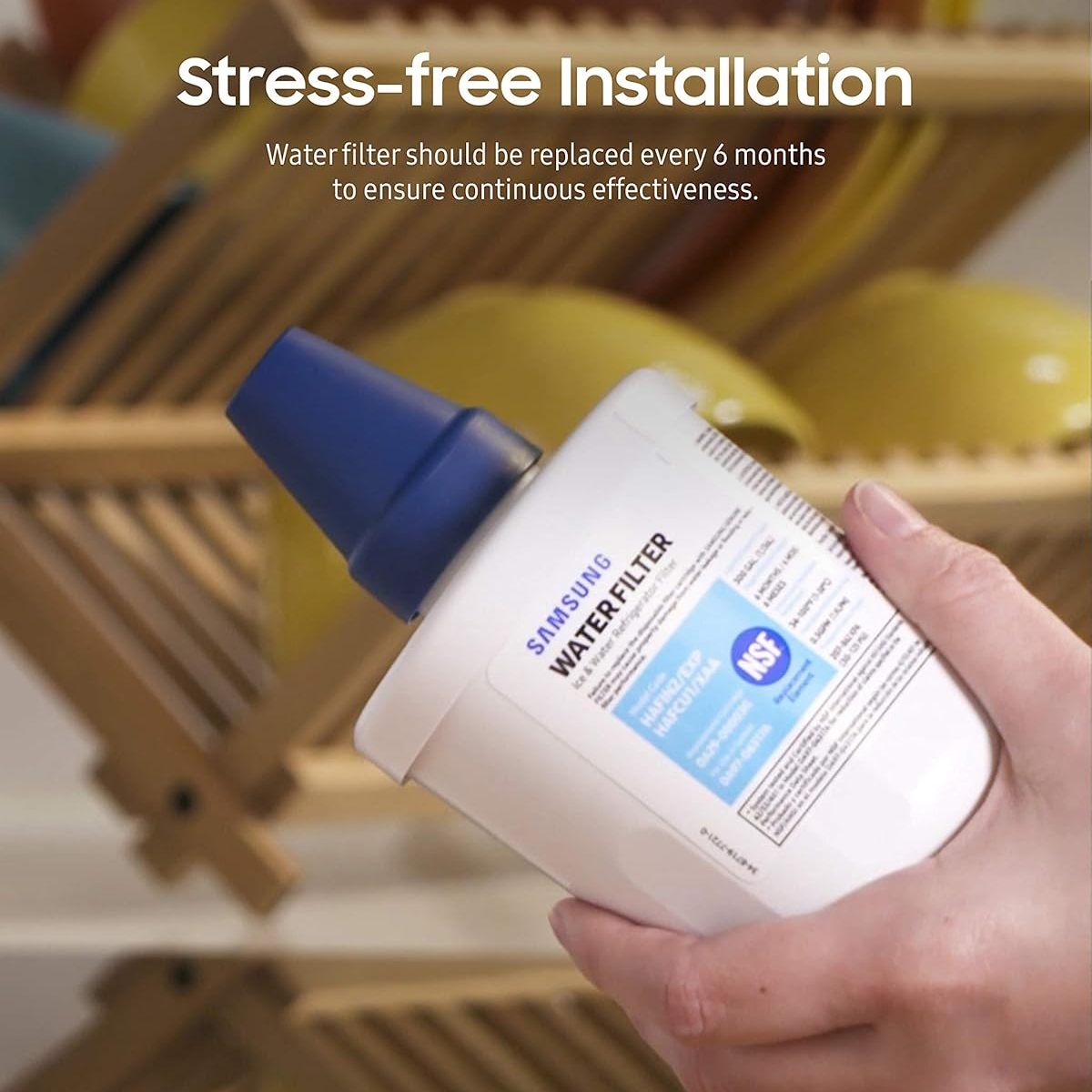 A hand holding a Samsung genuine water filter cartridge with a blue cap, set against a blurred background of wooden shelves and yellow bowls, with text overlay reading 'Stress-free Installation'.